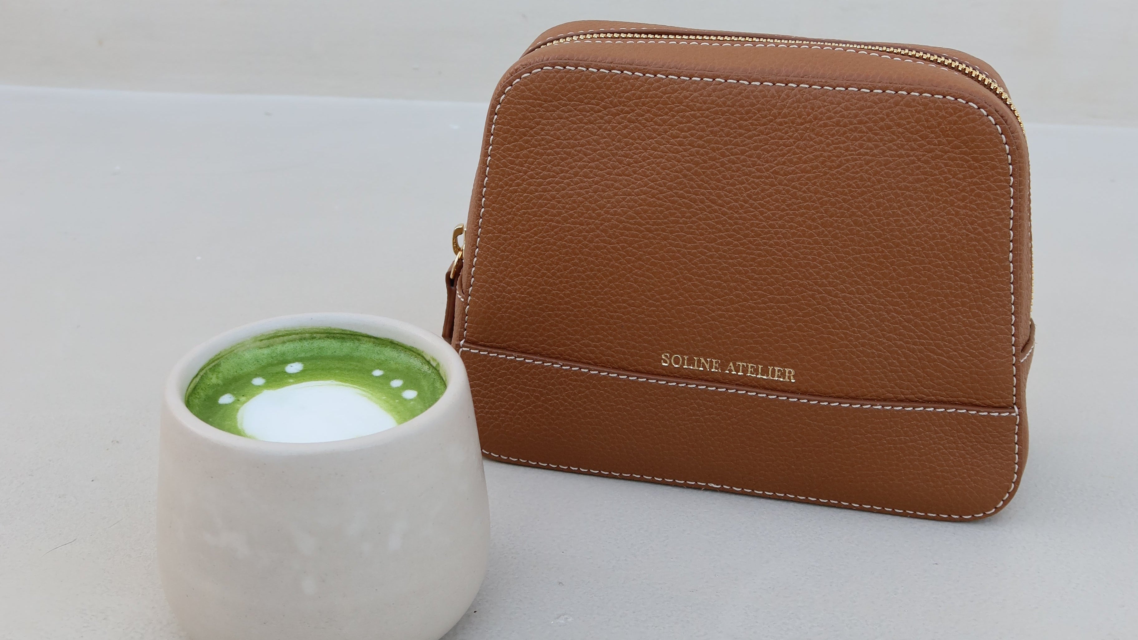 Soline Atelier EVERYDAY beauty case in brown leather with gold logo and white stitching next to a ceramic mug