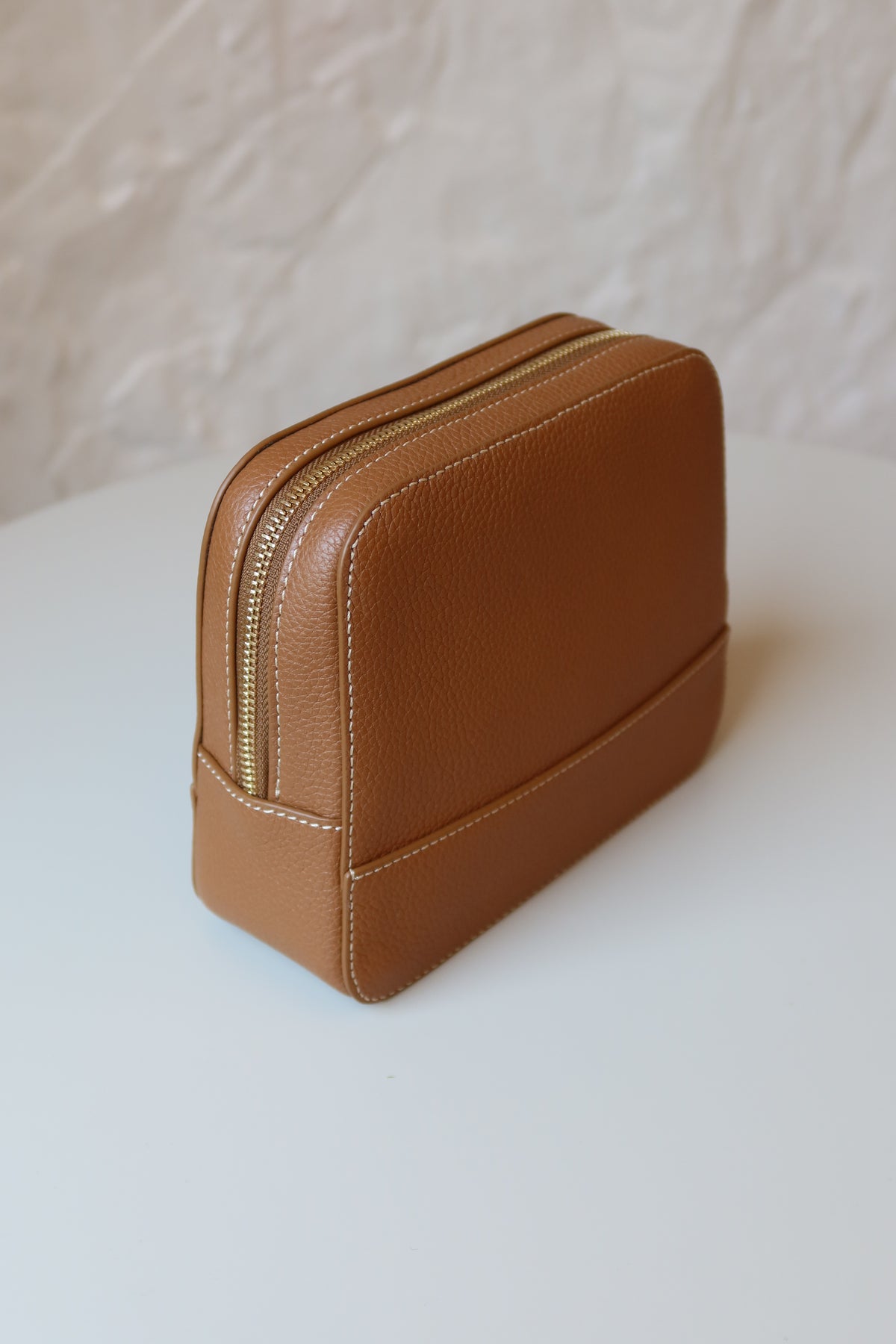 Soline Atelier EVERYDAY beauty case in camel leather with white stitching, gold logo and gold hardware standing on a white table, back view