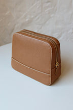 Soline Atelier EVERYDAY beauty case in camel leather with white stitching, gold logo and gold hardware standing on a white table, back view