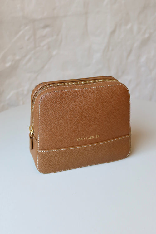 Soline Atelier EVERYDAY beauty case in camel leather with white stitching, gold logo and gold hardware on a white table, front view