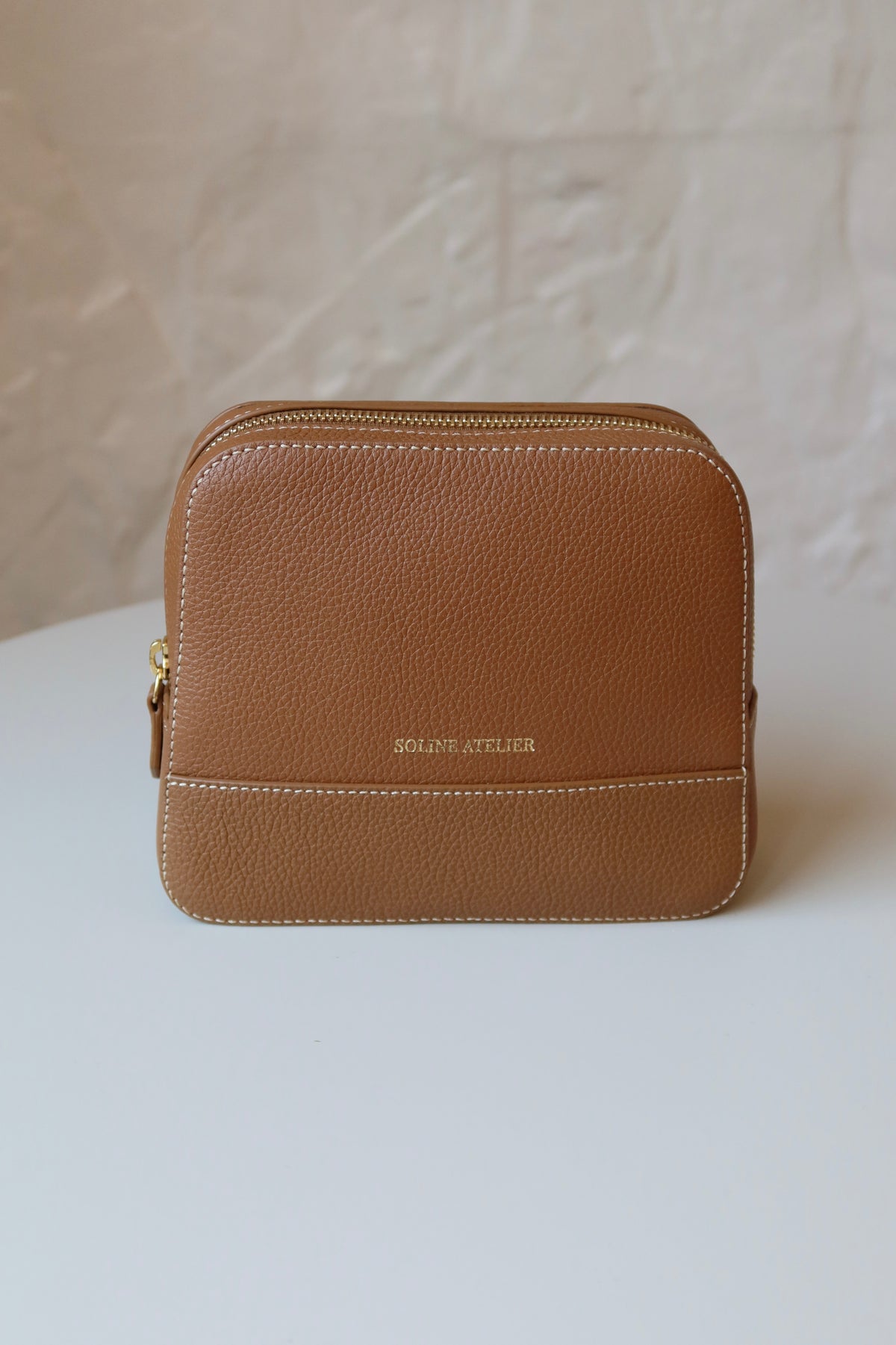 Soline Atelier EVERYDAY beauty case in camel leather with white stitching, gold logo and gold hardware standing on a white table, front view