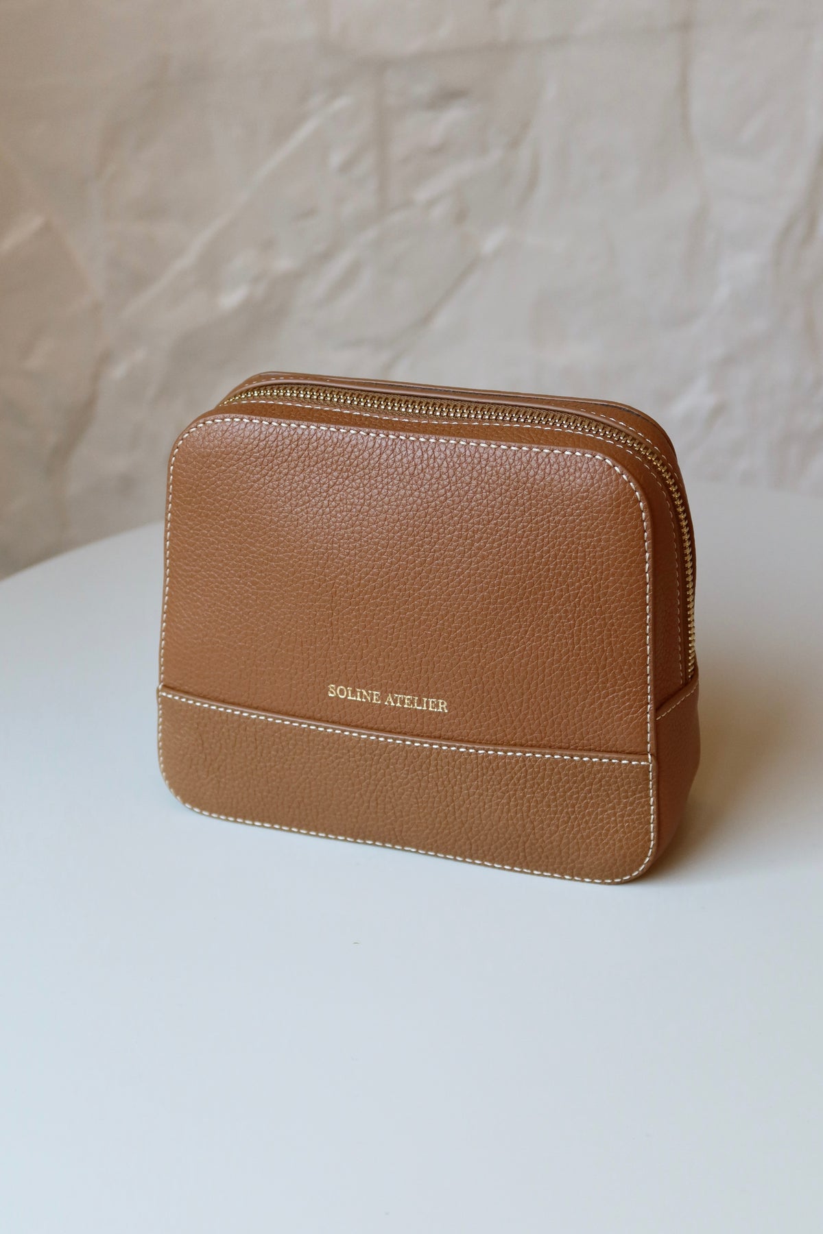 Soline Atelier EVERYDAY beauty case in camel leather with white stitching, gold logo and gold hardware on a white table, front view