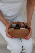 Woman in a white dress showing the inside of the Soline Atelier EVERYDAY beauty case in camel leather with white stitching, gold logo and gold hardware while sitting on a beige bench, top back view