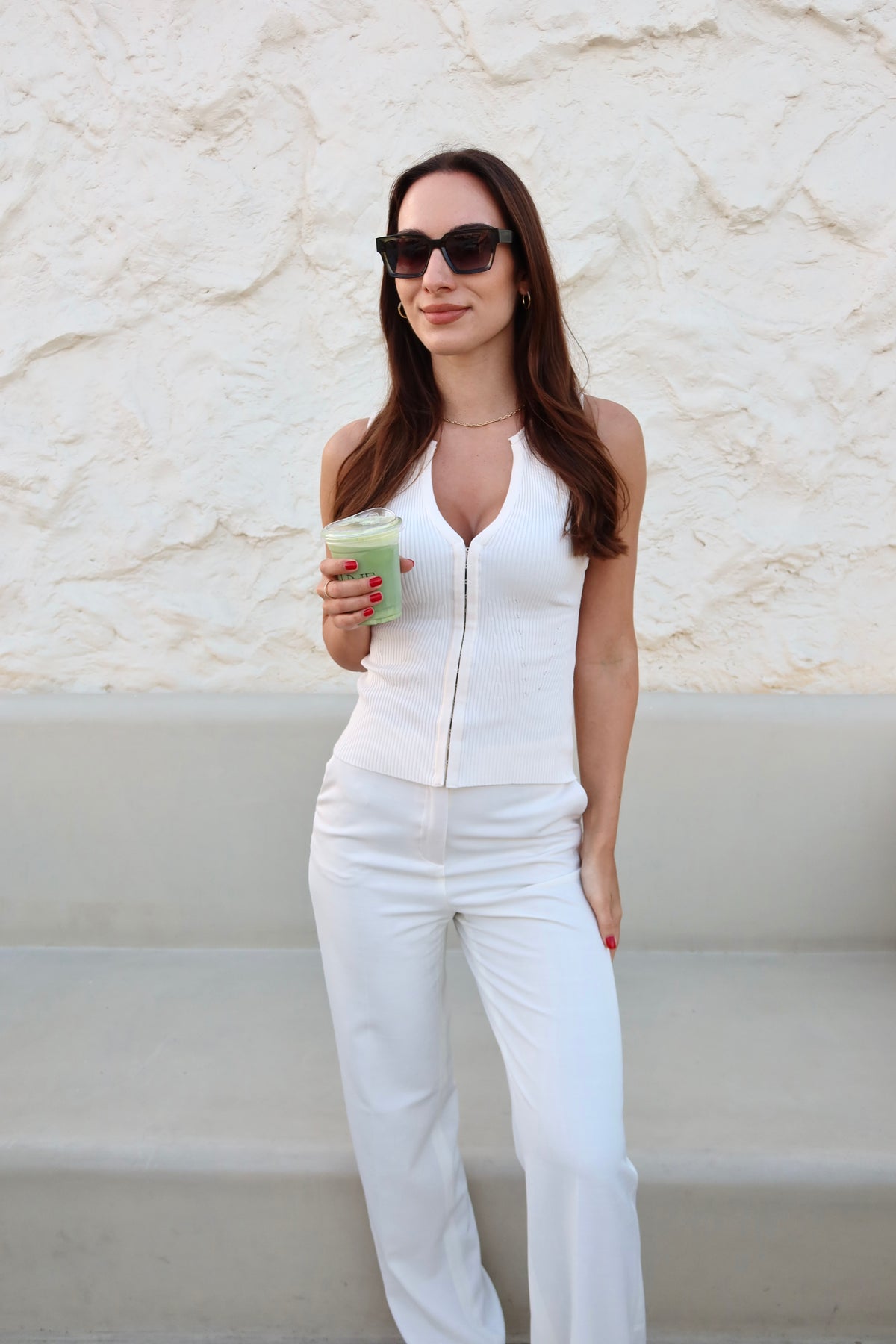 Woman wearing sunglasses in a white outfit holding a matcha cup in front of a beige bench and white wall