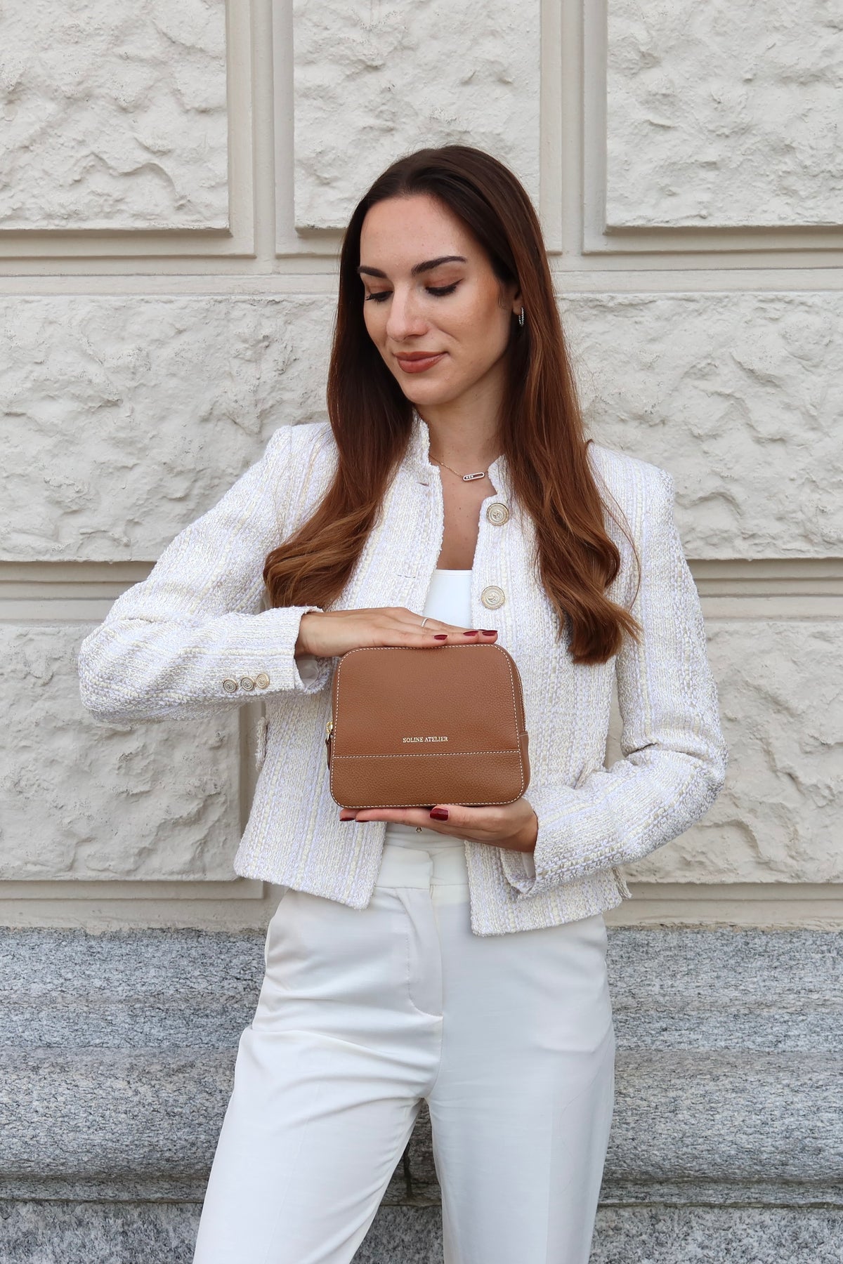 Woman in a white blazer and white trousers holding the Soline Atelier EVERYDAY beauty case in camel leather with white stitching, gold logo and gold hardware in front of a beige and grey wall